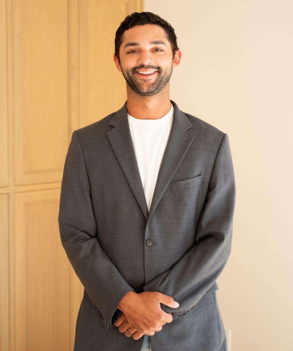 A man with short dark hair and a beard, wearing a gray suit over a white t-shirt, stands indoors in front of light wooden cabinets, smiling with his hands folded in front of him.