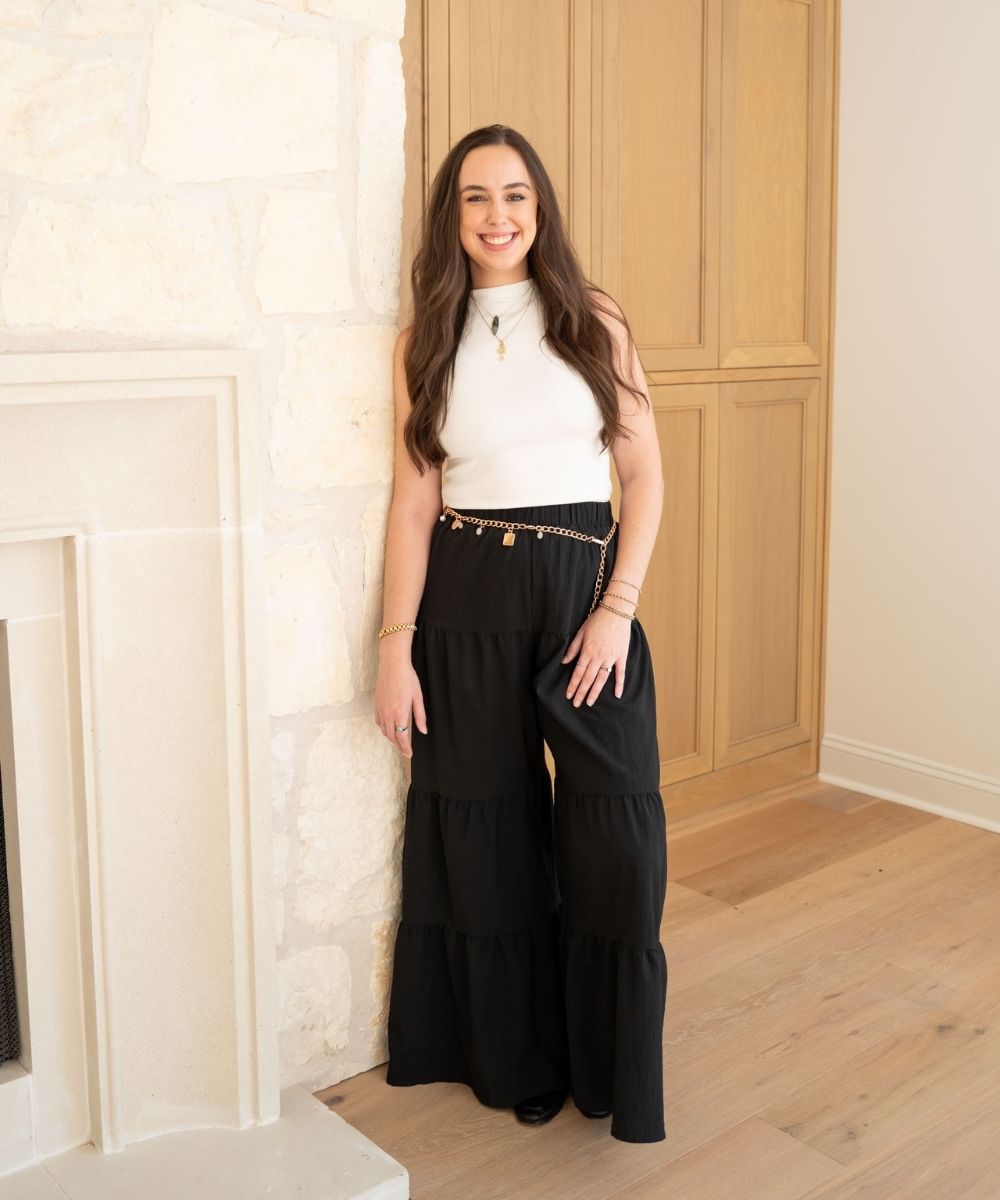 A woman with long brown hair stands indoors, smiling. She wears a sleeveless white top, black wide-leg pants with a chain belt, and gold jewelry. She is leaning against a light stone fireplace in a bright room with wood floors.