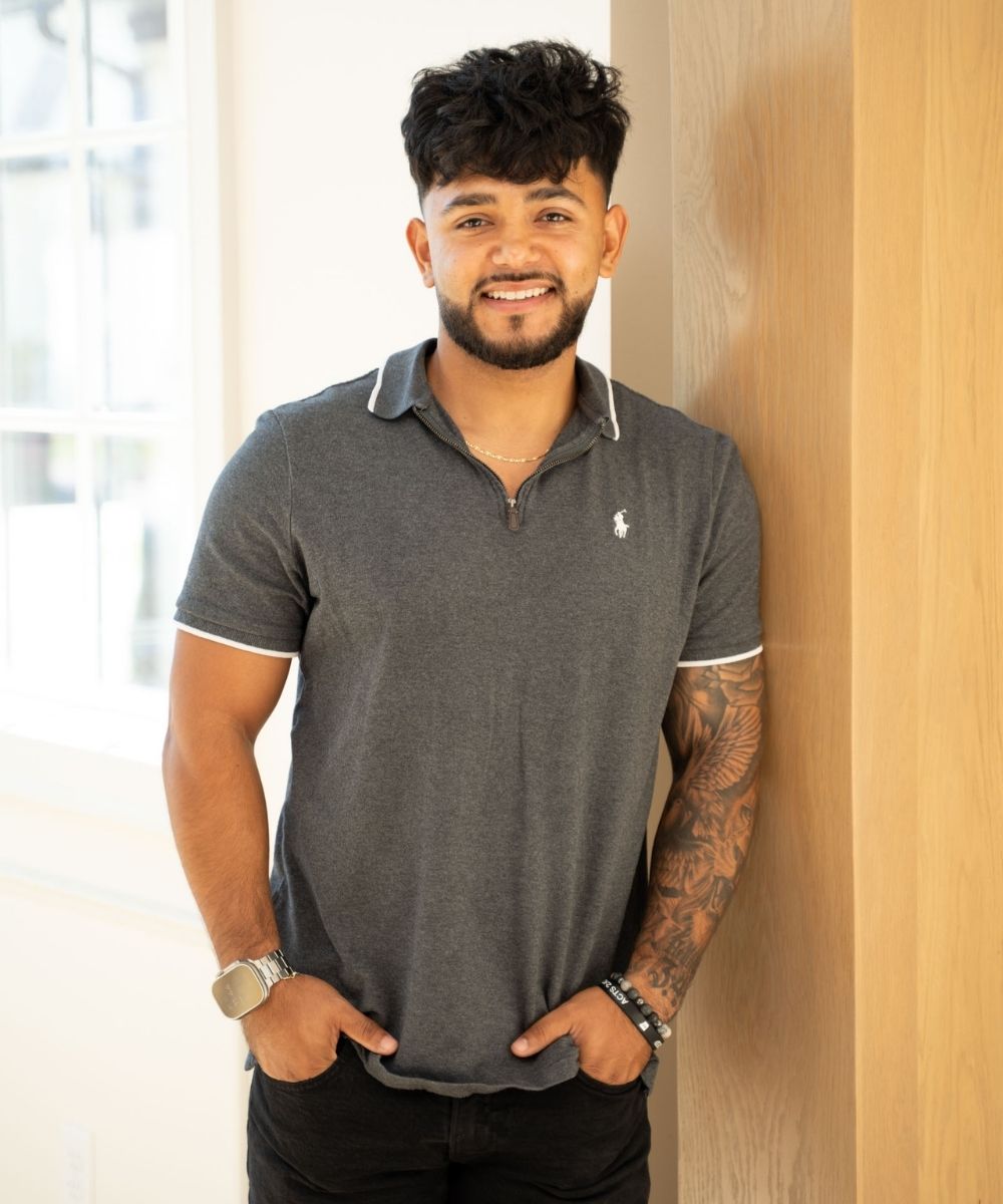 A smiling man with short, dark hair and a trimmed beard stands indoors, leaning against a light wooden wall. He wears a dark gray polo shirt, black pants, and has tattoos on his left arm. His hands are in his pockets.
