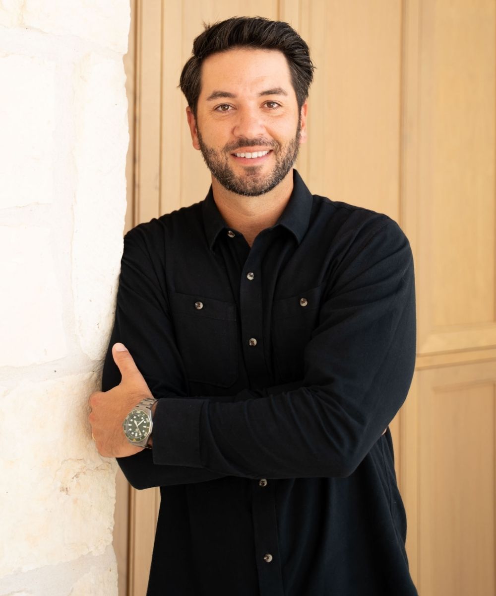 A man with dark hair and a beard, wearing a black button-up shirt and a watch, stands indoors with arms crossed, smiling and leaning against a light-colored stone wall.