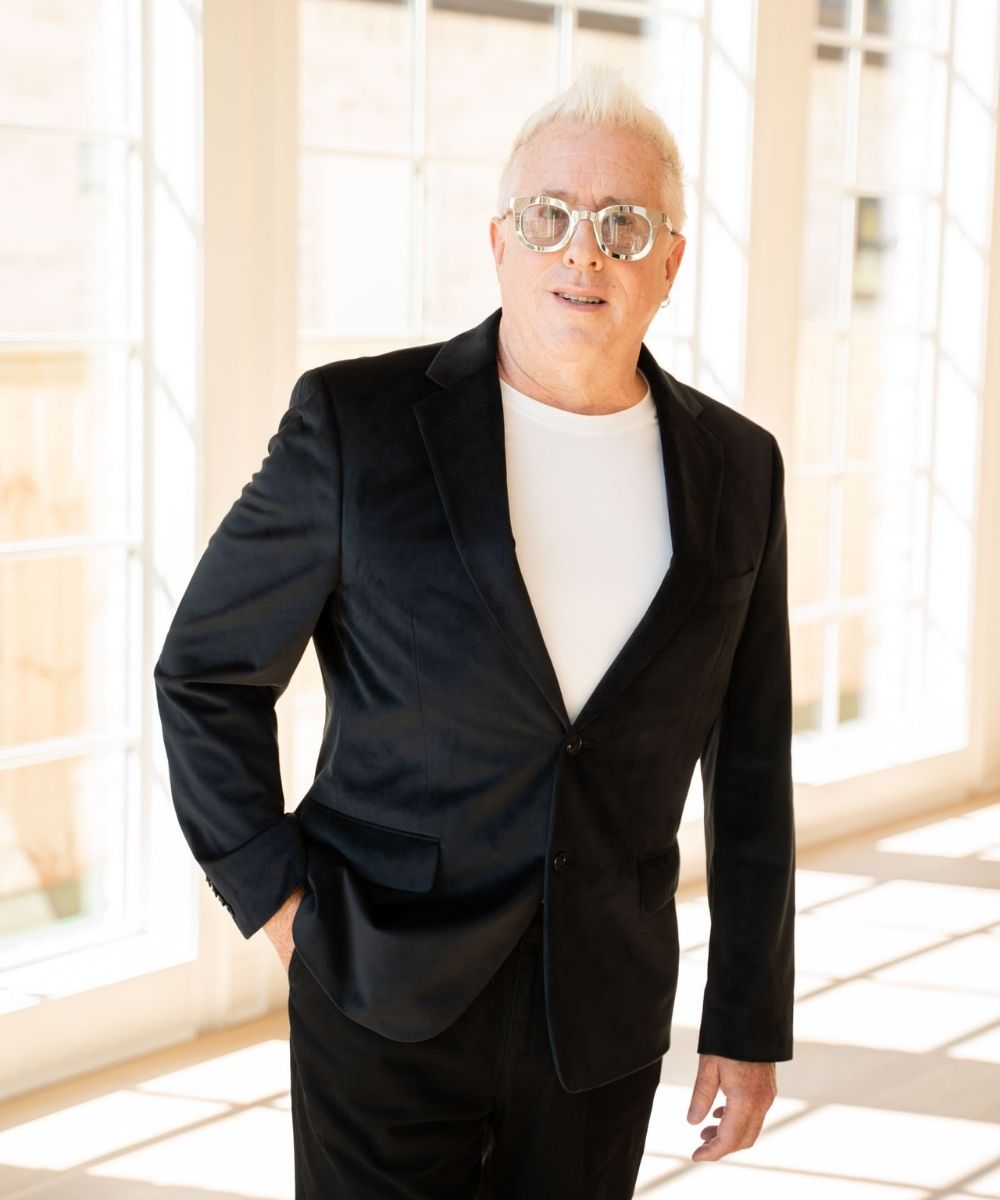 An older man with short white hair and glasses stands indoors by large windows, wearing a black suit jacket over a white shirt and smiling at the camera.