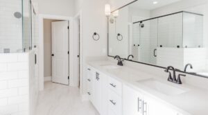 Bright, modern bathroom with white cabinets, double sinks, black fixtures, large mirrors, glass shower enclosure, white tiled walls, and a white door in the background. The space is clean and minimalist.