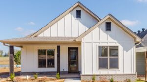 A modern farmhouse-style home with white vertical siding, a peaked roof, brick trim along the bottom, large front windows, and a small landscaped garden in front under a clear blue sky.
