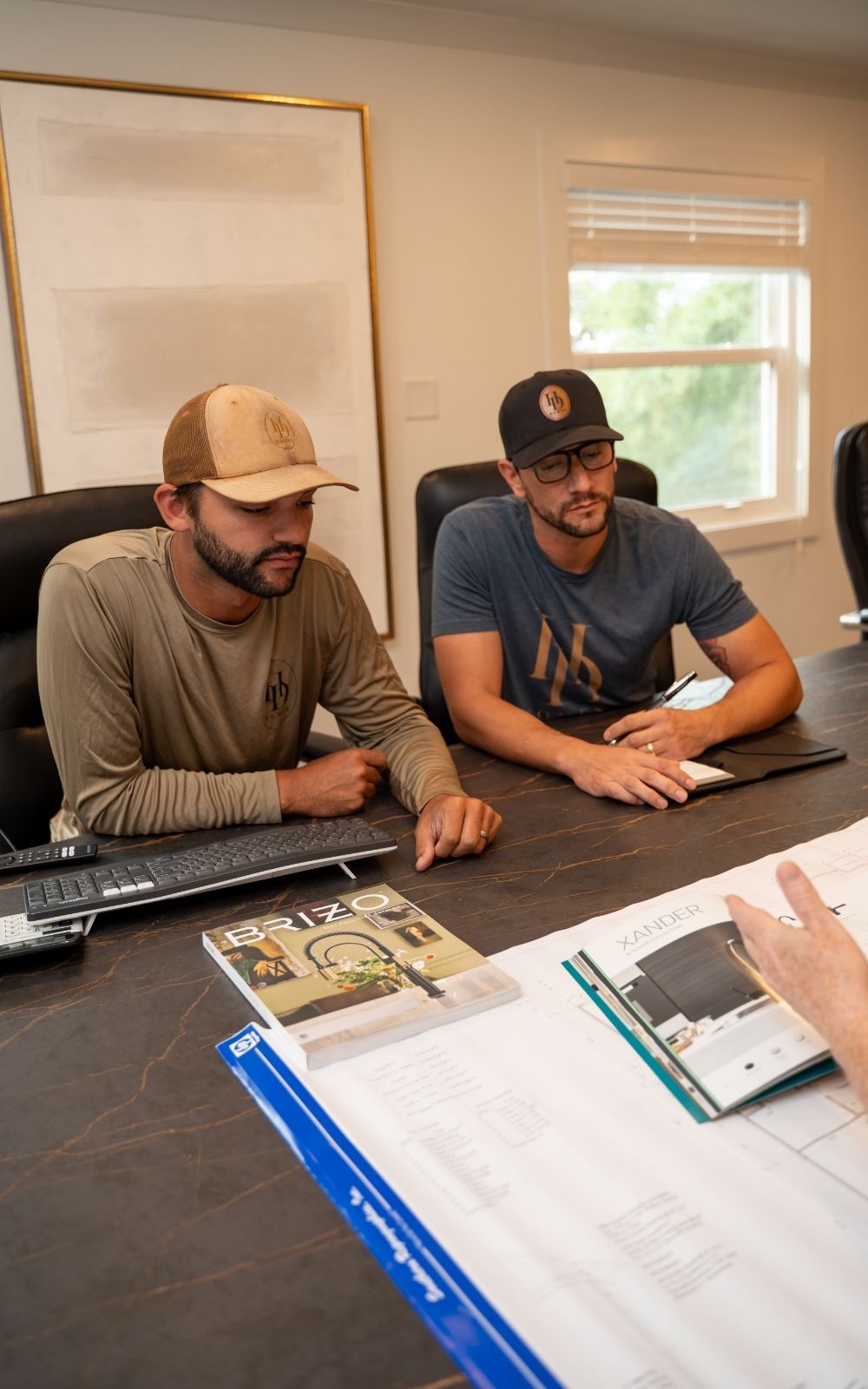 Two men in casual attire sit at a desk, looking at documents and a computer keyboard, while a third person gestures toward brochures and papers spread on the table.