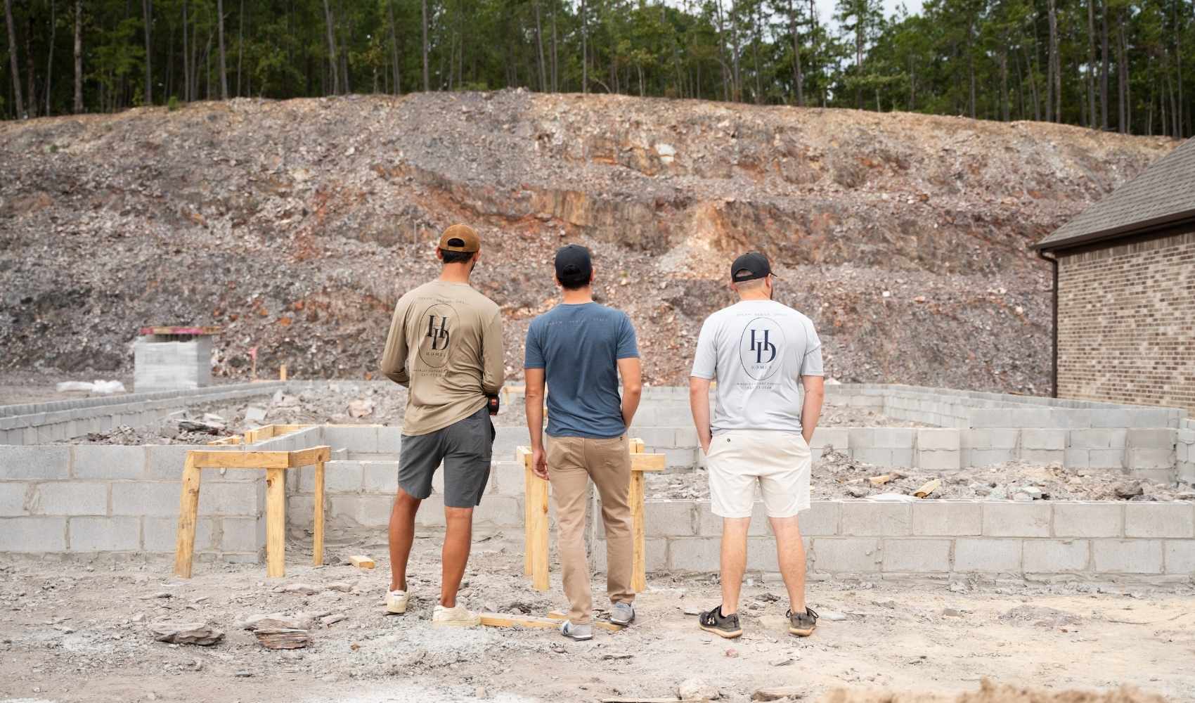 Three men stand with their backs to the camera, observing the foundation of a building under construction in a rocky, partially cleared area with trees in the background.