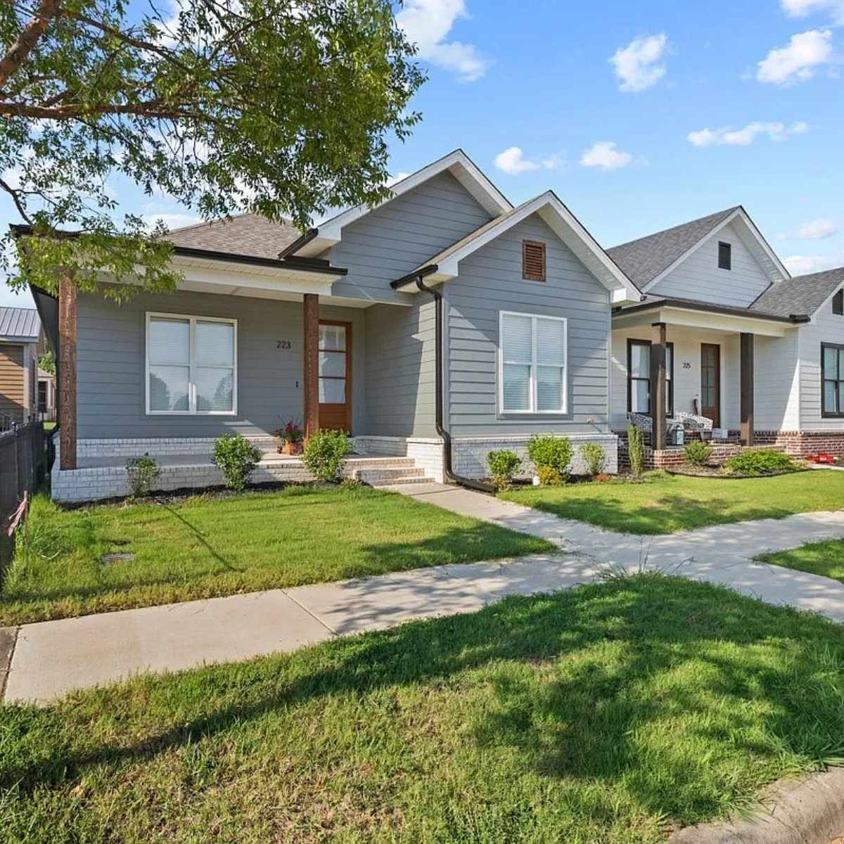 A modern gray house with white trim, a covered porch, wooden columns, and a well-kept lawn. The house has large front windows, brick accents, and is situated on a tree-lined sidewalk in a suburban neighborhood.