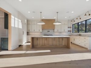 Spacious modern kitchen with light wood floors, a large central island, white cabinets, stainless steel appliances, pendant lights, and a big window letting in natural light.