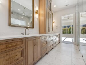 Modern bathroom with light wood cabinets, double sinks with gold hardware, large mirrors, marble countertops, and white marble floor tiles. Glass shower on the right and a bright room visible through glass doors at the back.