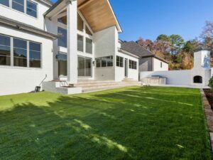 Backyard with artificial grass, a modern white house featuring large windows, a covered patio with wood ceiling, steps leading to the yard, an outdoor kitchen, and a white stone fireplace against a fenced wall.