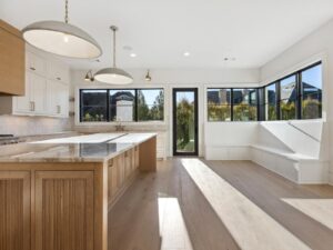 Bright, modern kitchen with large island, pendant lights, light wood floors, white cabinets, and a built-in bench by large black-framed windows. Glass door leads outside, filling the space with natural light.