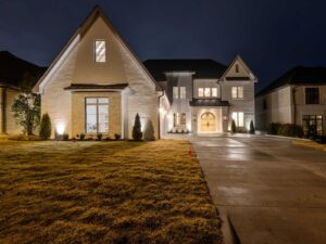 A large, modern two-story house with white brick exterior and multiple windows is brightly lit at night. The driveway and front lawn are visible in the foreground.