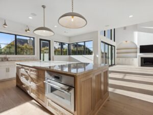 Modern, bright kitchen with wooden island, marble countertop, built-in stainless steel oven, white cabinets, pendant lights, and large windows letting in natural light; open living area with fireplace in the background.