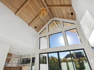 A modern living space featuring a vaulted wooden ceiling with a chandelier, large floor-to-ceiling windows letting in natural light, and an open view of a kitchen with wood accents.