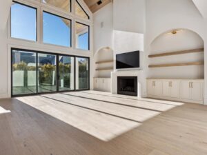 Bright, modern living room with large windows, glass doors, light wood flooring, built-in shelves, cabinets, a fireplace, and a wall-mounted TV. Sunlight streams in, creating a spacious, airy feel.