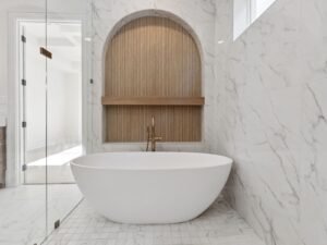 Modern bathroom with a white oval bathtub, marble walls, and a built-in wood accent alcove with a gold faucet. Natural light enters through a high window and an open glass door.