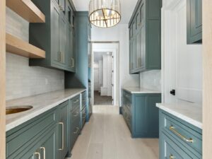 A modern kitchen pantry with green cabinets, gold handles, white countertops, open wooden shelves, light wood flooring, and a round chandelier. A doorway leads to another room in the background.