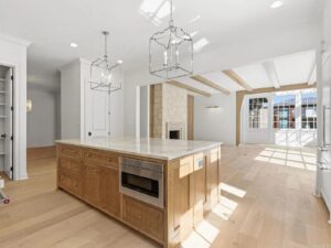 Bright, modern kitchen with light wood floors, a large island with built-in microwave, pendant lights, and open living area featuring a stone fireplace and exposed ceiling beams. Large windows let in ample natural light.