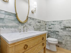 A bathroom with a wooden vanity and marble countertop, an oval mirror with a gold frame, wall-mounted light fixture, green and gray tile backsplash, and a white toilet.