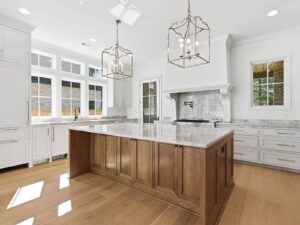 A modern kitchen with white cabinets, a large wooden island with a marble countertop, light wood floors, two chandeliers, large windows, and stainless steel appliances.