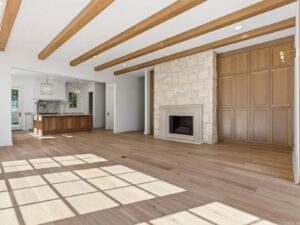 Bright living room with light wood floors, exposed wooden ceiling beams, a stone fireplace, large built-in wooden cabinets, and open layout connecting to a modern kitchen with pendant lights. Sunlight streams through large windows.