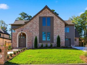 A two-story brick house with tall windows, arched wooden front door, landscaped front yard, and driveway, set against a background of green trees and a clear blue sky.