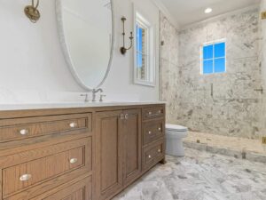 A modern bathroom with a large wood vanity, oval mirror, marble countertop, and a walk-in marble shower with a window. The space features neutral tones and elegant fixtures.