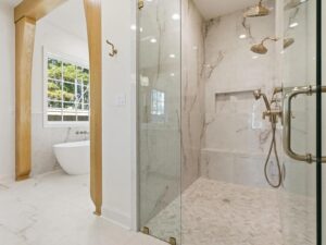 A modern bathroom with a large glass-enclosed shower, marble walls, dual shower heads, a built-in bench, and a freestanding bathtub near a window with natural light.