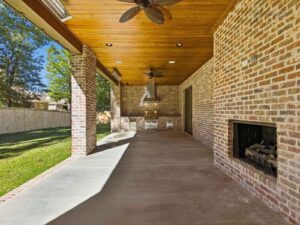 Covered patio with a wooden ceiling, ceiling fans, and brick walls, featuring an outdoor fireplace and built-in grill area. The space opens to a grassy backyard with a wooden fence and trees.
