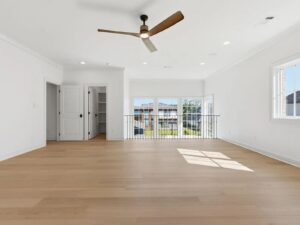 Spacious, bright room with light wood flooring, white walls, a ceiling fan, and large windows letting in sunlight. Glass doors open to a balcony with a view of neighboring buildings.