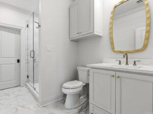 A modern bathroom with white marble floor, a glass shower, white cabinets, a toilet, and a vanity with a gold-framed mirror and matching gold faucet. The decor is clean and minimalist.