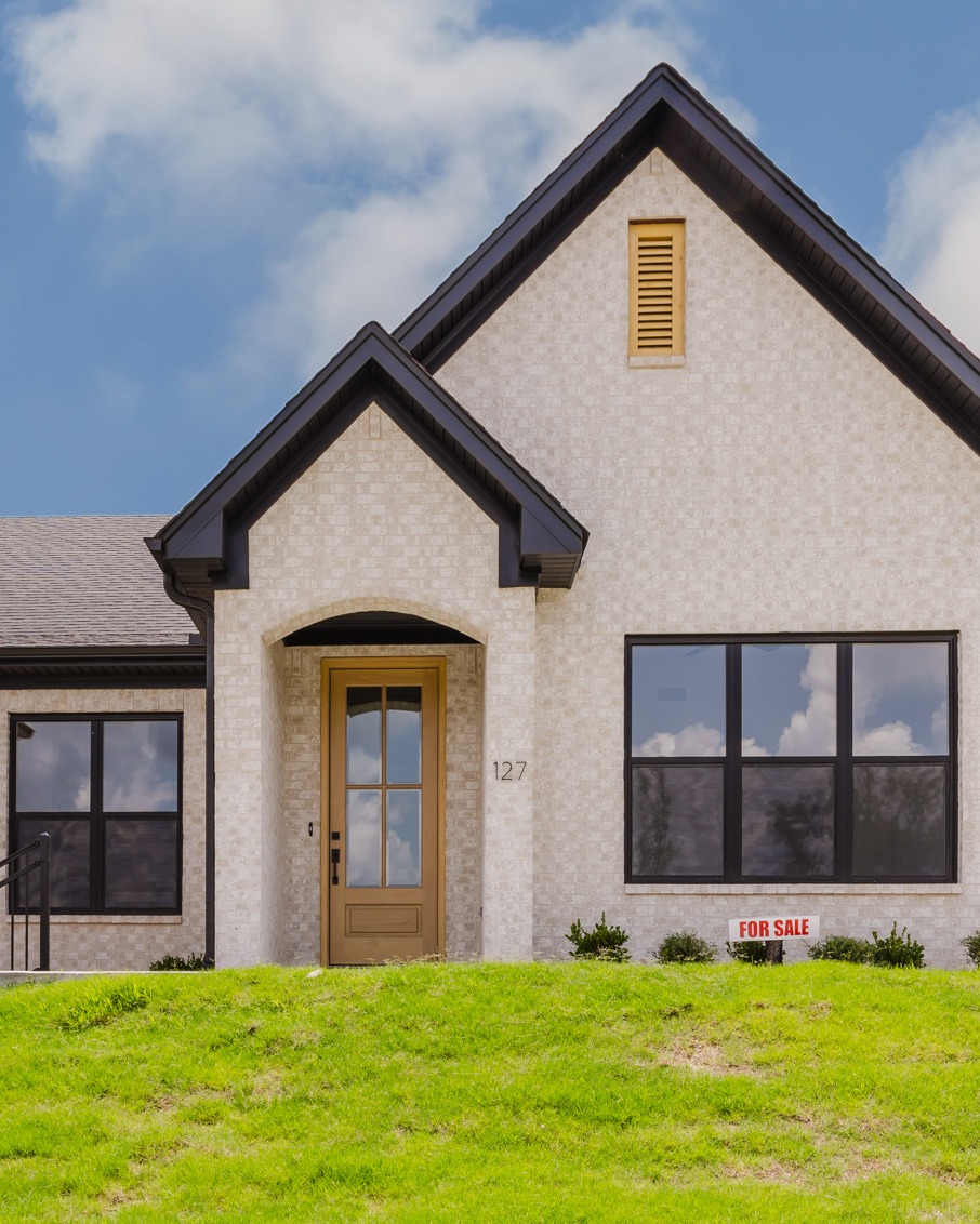 A modern, light brick house with large windows, a brown front door, and a well-kept green lawn. A For Sale sign is visible in front of the house, and the sky is partly cloudy.