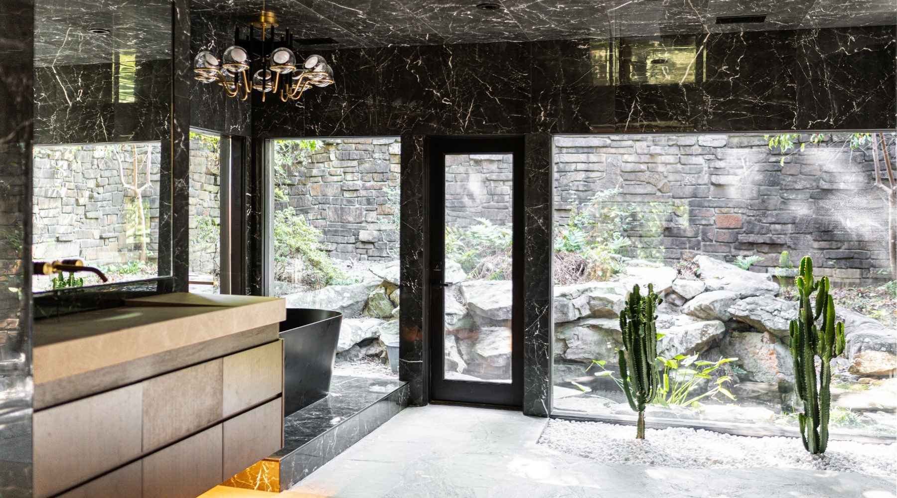 Modern bathroom with black marble walls and floor, large windows showing a stone garden outside, minimalistic vanity, black bathtub, chandelier, and two tall cacti in a white gravel indoor garden.