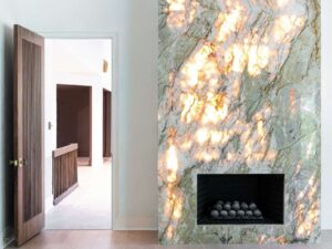 A modern fireplace with a glowing, backlit marble facade featuring warm, golden patterns stands next to an open wooden door leading to a bright hallway with dark railing and white walls.