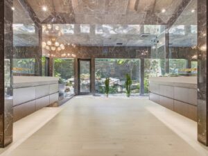 Modern bathroom with marble walls and ceiling, two large vanities, bright floor, and large mirrors reflecting natural light from a glass door and windows overlooking a lush garden.