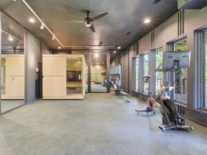 Modern gym room with large windows, exercise bikes, a wall-mounted TV, mirrors, and a wooden sauna or steam room; bright and spacious with natural light.