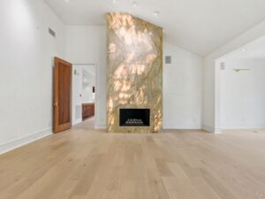 A modern, empty living room with light wood flooring, white walls, and a fireplace featuring a tall, polished stone surround with beige and gold veining. An open wooden door leads to another room.