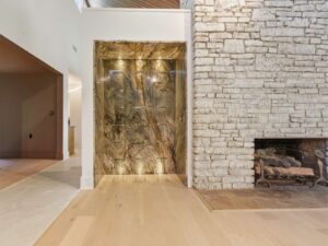 A modern living space featuring a white stone fireplace on the right and a built-in, backlit marble display shelf on the left, with light wood flooring and an open doorway to adjoining rooms.