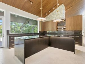 Modern kitchen with black marble countertops, wood cabinets, brass hardware, pendant lights, a slanted wood ceiling, large windows, and a stone accent wall with a built-in oven. Natural light fills the spacious area.