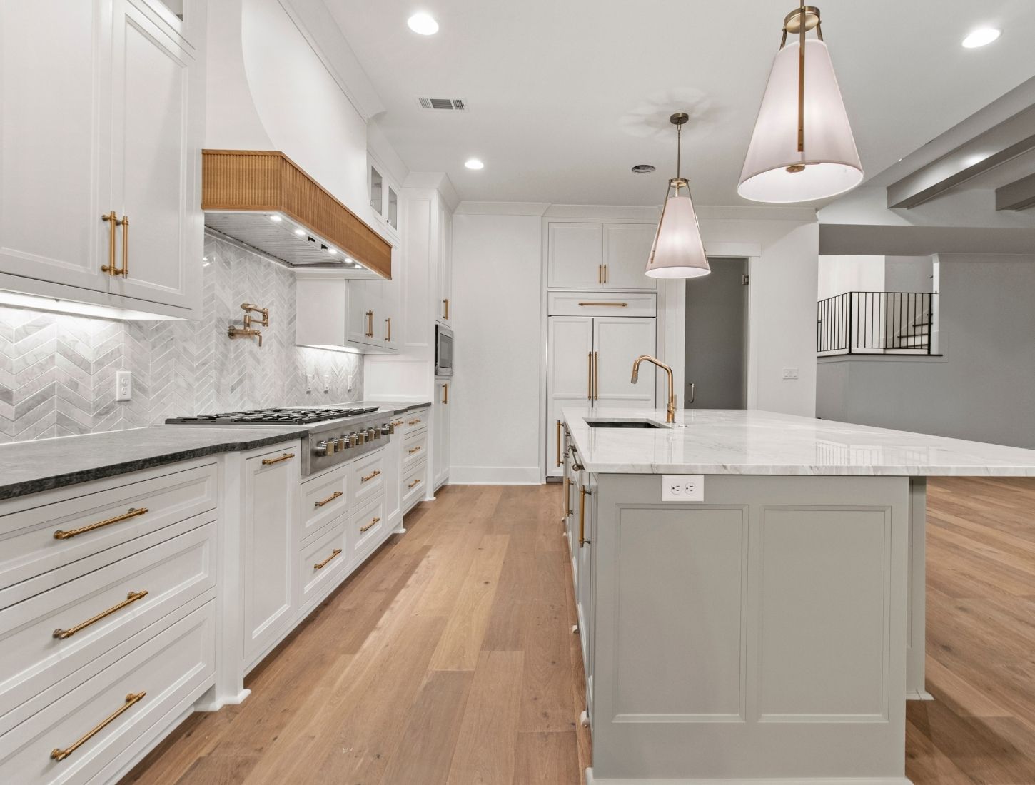 Modern kitchen with white cabinets, stainless steel appliances, a large island with a marble countertop, gold fixtures, pendant lights, and light wood flooring. Herringbone backsplash and open shelving above the stove.