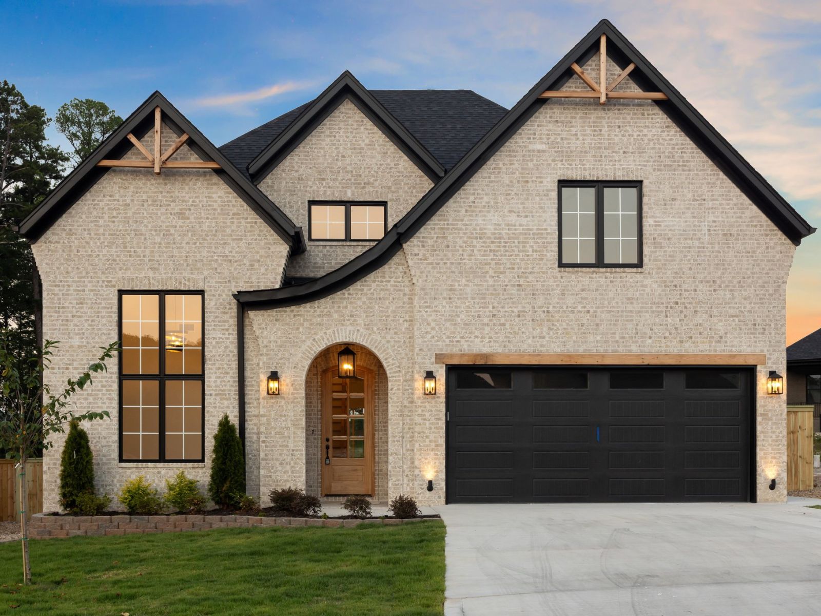 Modern two-story brick house with dark roof and garage doors, warm exterior lights, large windows, and landscaped front yard with a small tree and green grass at sunset.