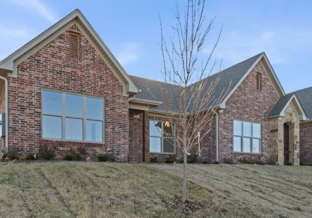 Single-story brick house with multiple large windows, a front gable roof, and a small young tree on a sloped lawn; clear blue sky in the background.