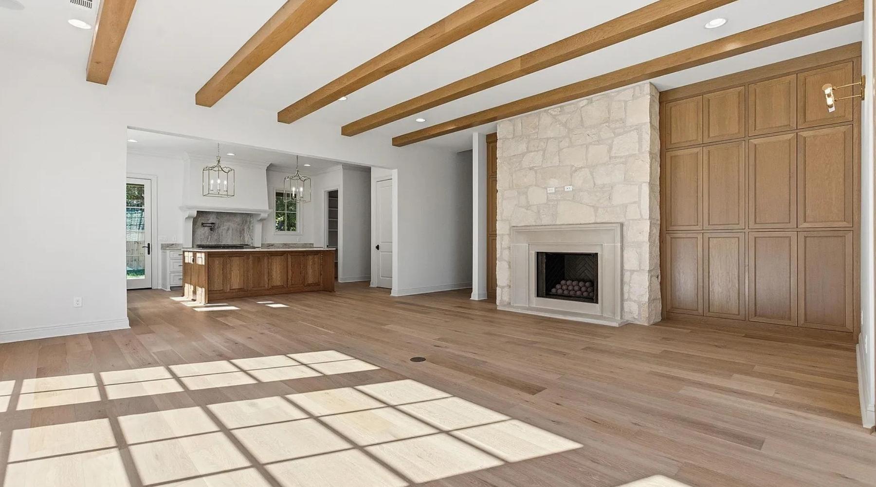 A bright, spacious living room with wooden ceiling beams, light wood floors, a stone fireplace, built-in wood cabinets, and an open view into a modern kitchen with an island and pendant lights. Sunlight streams in through large windows.