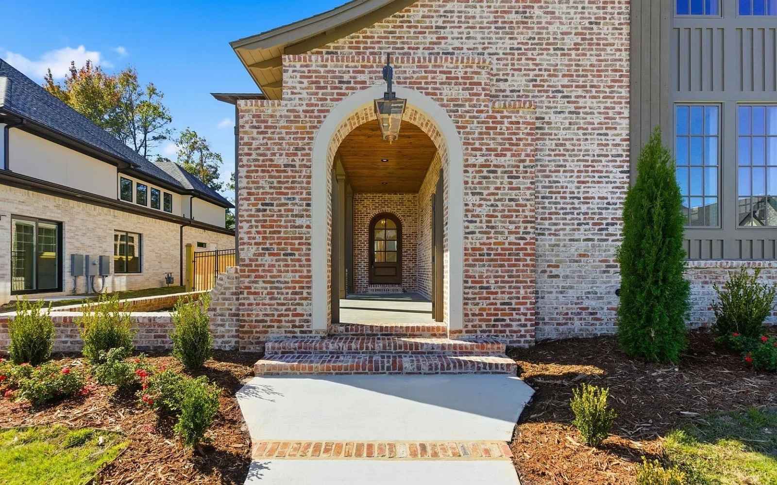 Brick house with an arched entryway, wooden ceiling, and lantern-style light fixture. A concrete path leads to the front door, surrounded by landscaped bushes and mulch. Neighboring homes are visible on the left.