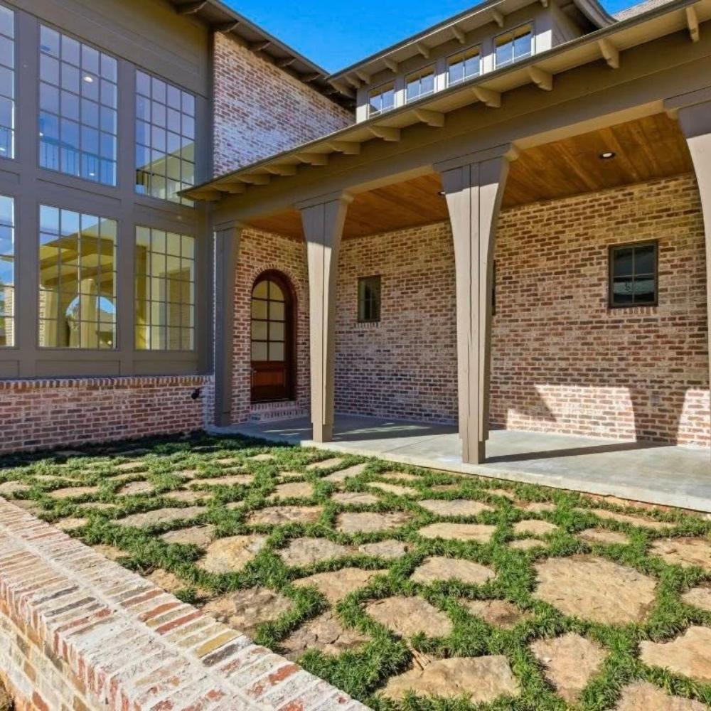 A brick house with tall windows and a covered entryway. The front yard features large stone pavers set in a geometric pattern with grass growing between them. Blue sky is visible above the house.
