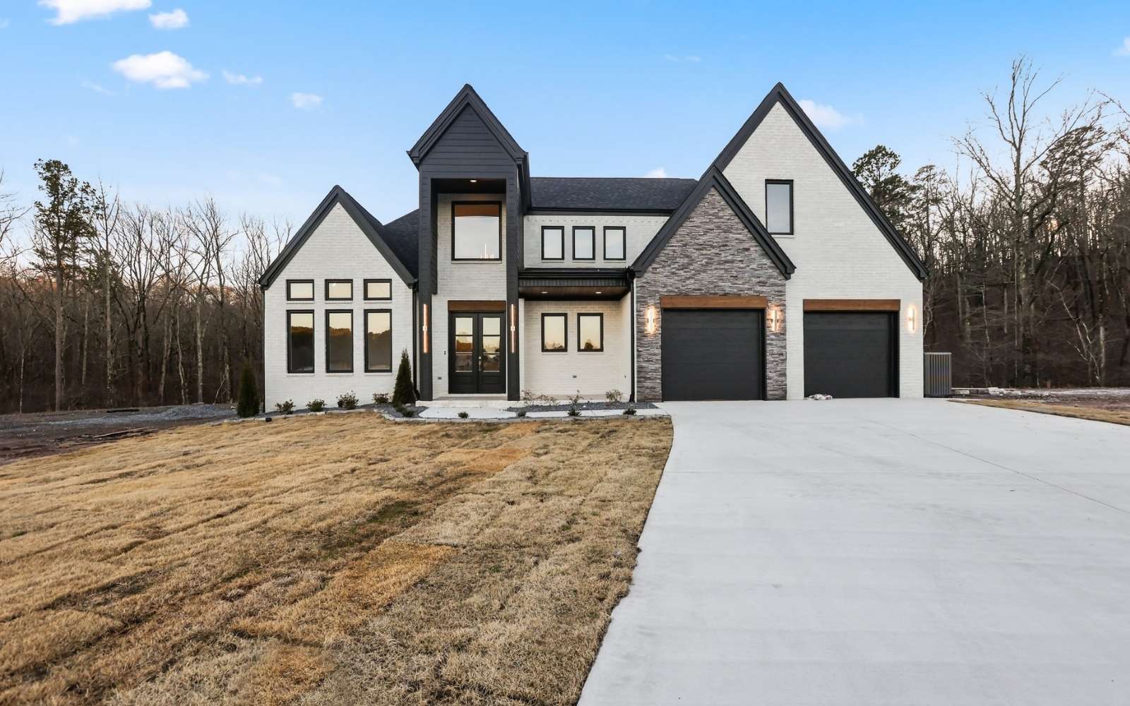 Modern two-story house with large windows, a stone and white exterior, and two-car garage, set on a spacious lot with a wide driveway and surrounded by trees.