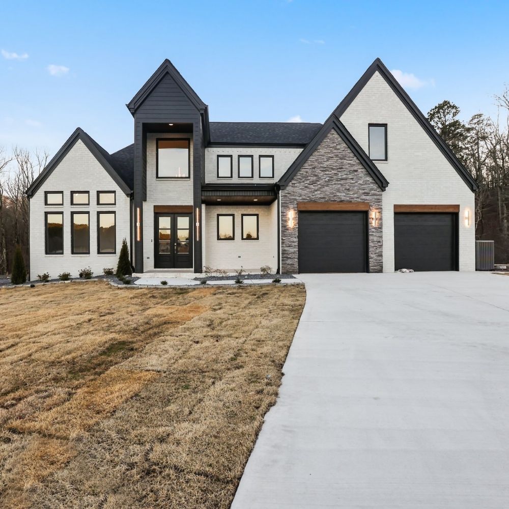 Modern two-story house with white and gray exterior, large windows, double garage, and a wide concrete driveway. The front yard has minimal landscaping and brown grass, with trees in the background under a clear sky.