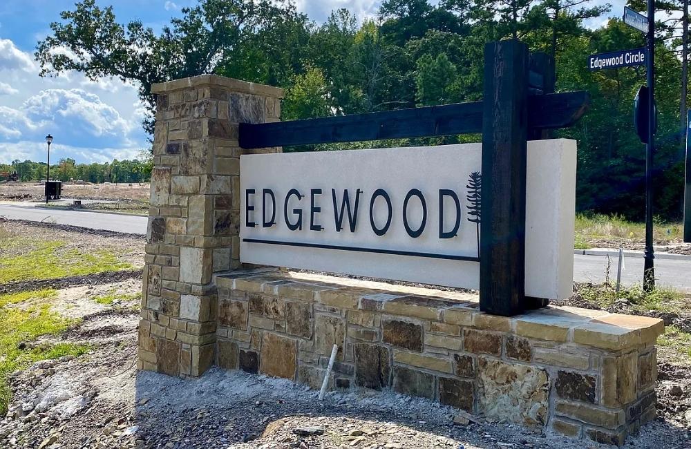 A stone and wood sign at a street corner displays the word EDGEWOOD with a small fern graphic; a blue street sign above reads Edgewood Circle. Trees and a partly cloudy sky are in the background.