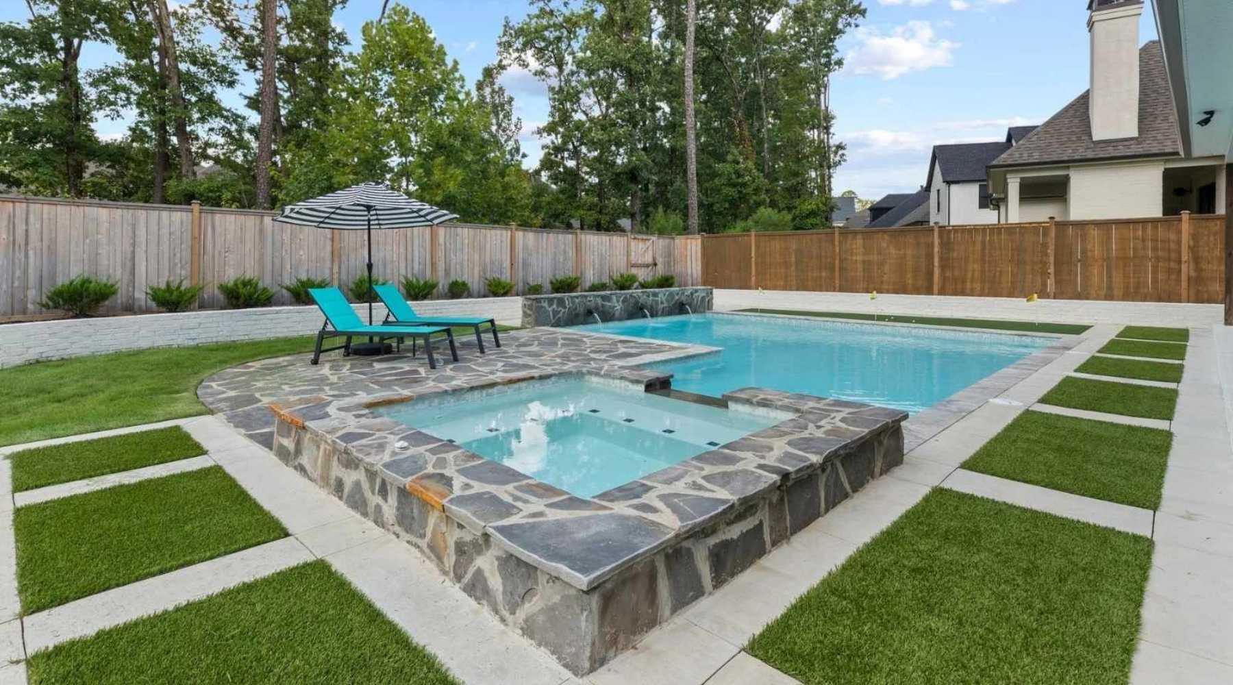 Modern backyard with a stone-edged pool and attached hot tub, two turquoise lounge chairs under a striped umbrella, green grass, paved walkways, and a wooden privacy fence surrounded by trees.