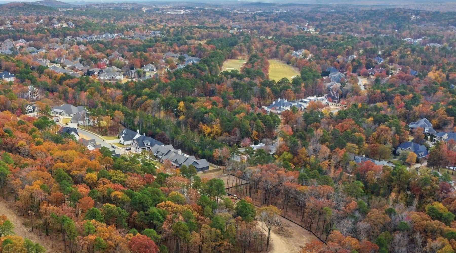 Aerial view of a suburban neighborhood surrounded by dense forest with autumn foliage in shades of orange, red, and green, with houses and winding roads scattered throughout the landscape.