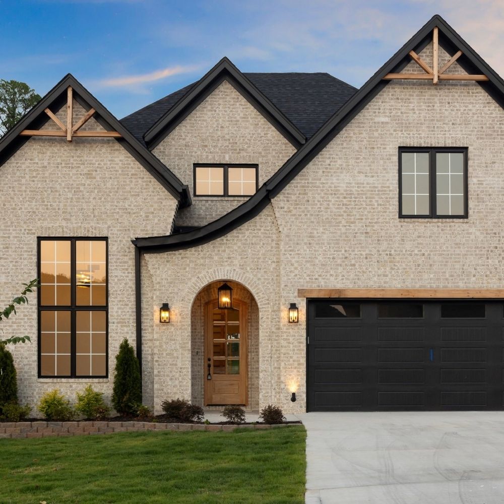 Modern two-story brick house with black trim, wooden accents, a double garage door, large windows, and a wooden front door with warm lights on either side, set against a landscaped yard at dusk.